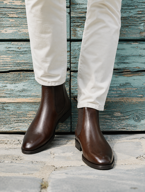 A person wearing dark brown leather Chelsea boots with elastic side panels and white trousers, standing against a weathered blue-green wooden plank wall.