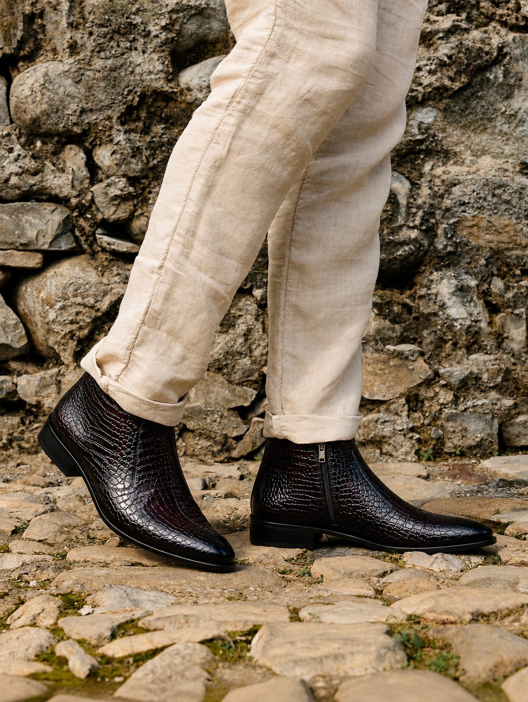 A person wearing black crocodile-embossed leather ankle boots with beige linen trousers, standing on a rustic stone path against a stone wall.