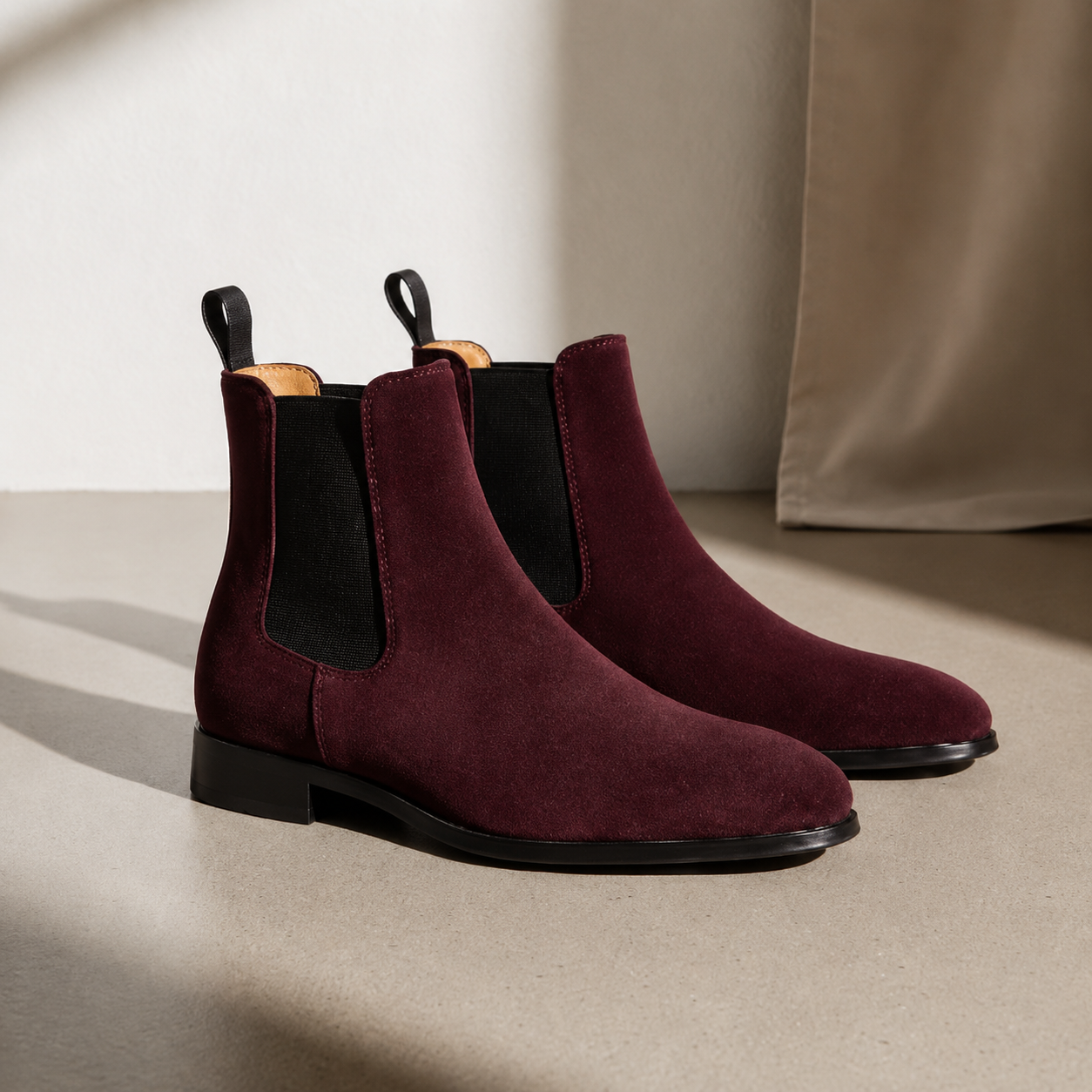 Pair of maroon Chelsea boots on a beige surface with a neutral background