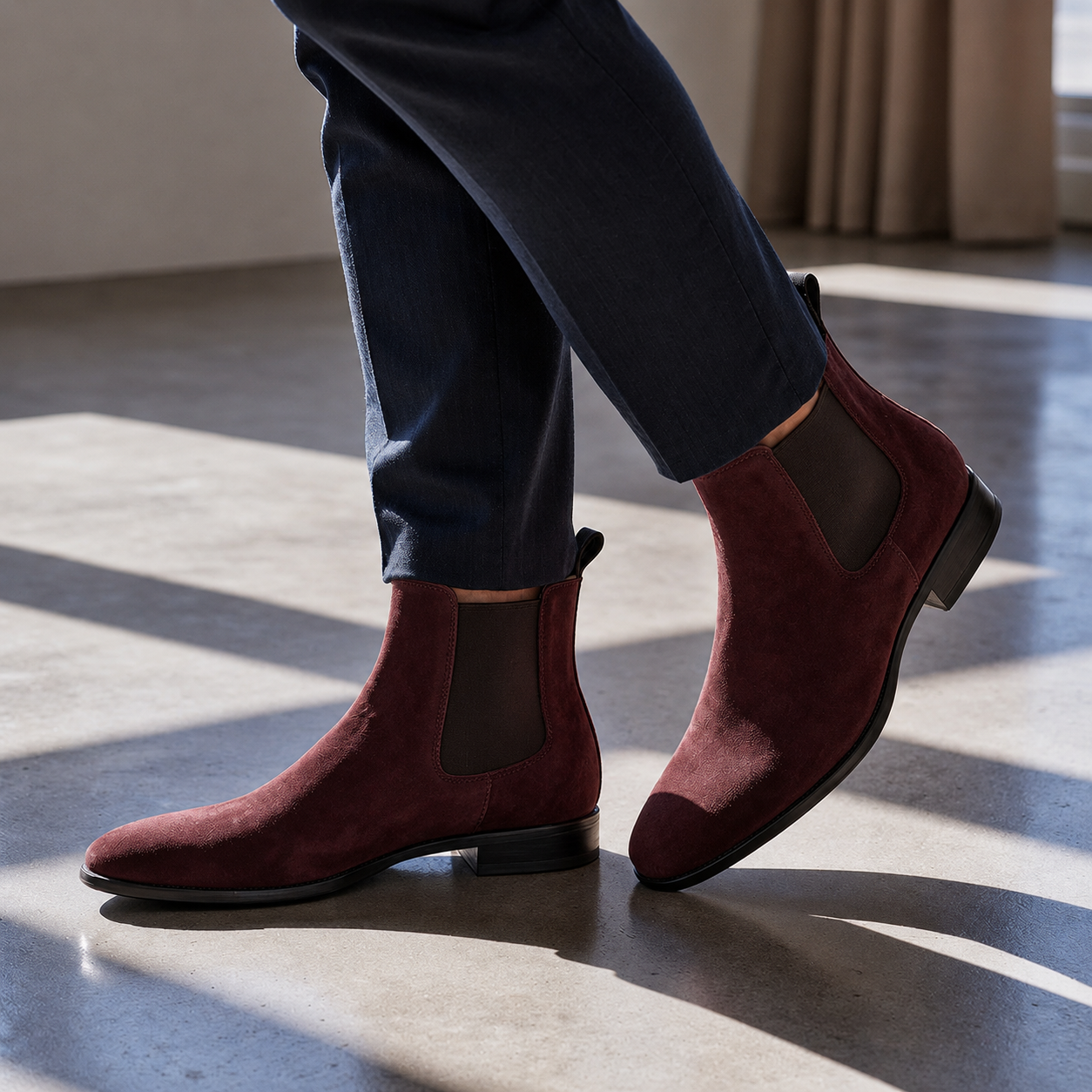 Red suede Chelsea boots worn with dark pants on a reflective floor.