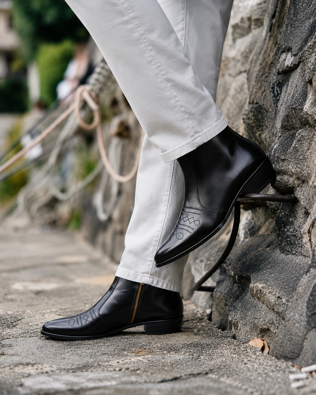 A close-up lifestyle shot of a person wearing black leather ankle boots with decorative stitching and white trousers, standing on a stone path next to a rustic stone wall.