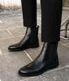 A pair of black leather Chelsea boots worn with black trousers on a gray stone pavement.