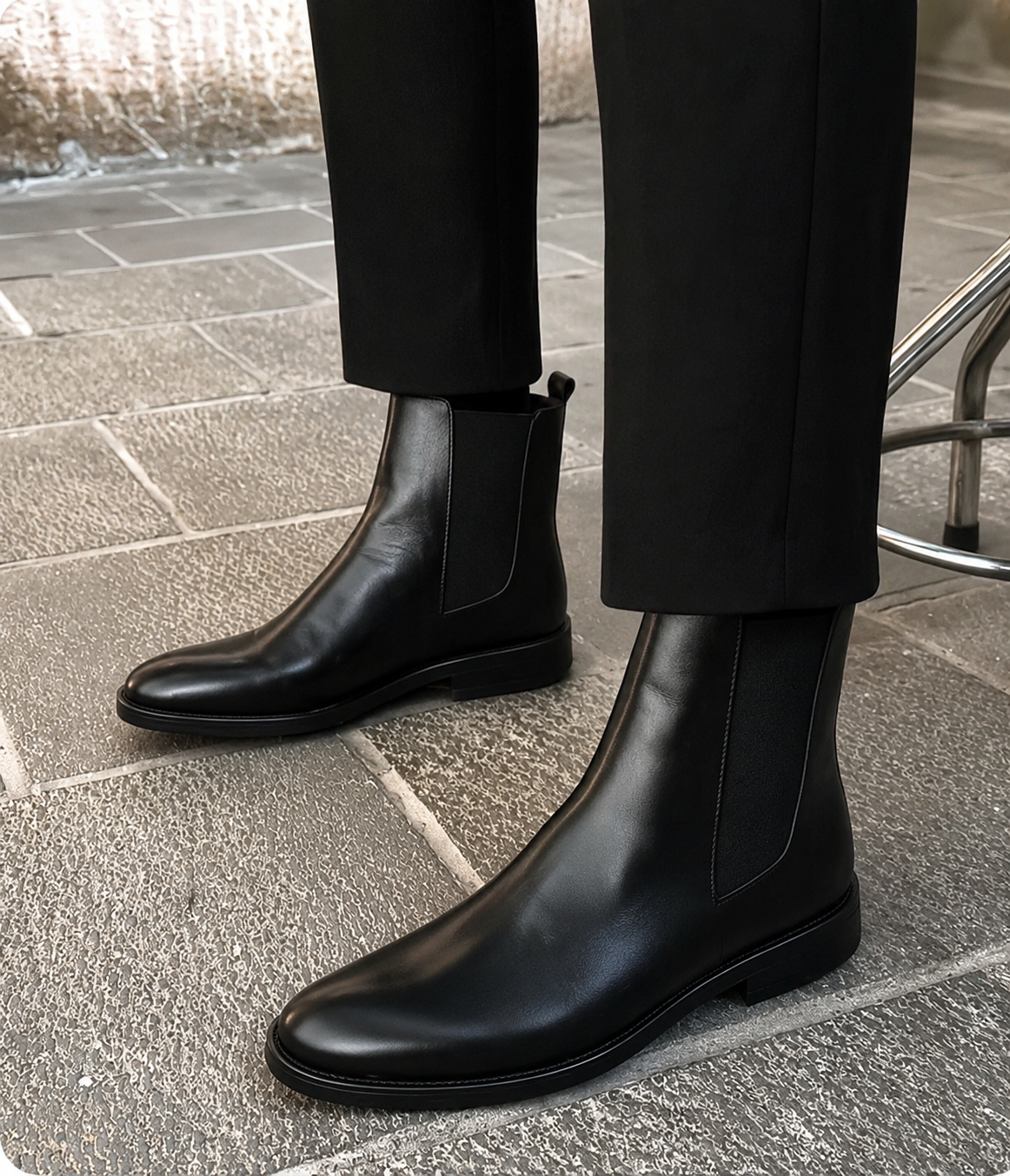 A pair of black leather Chelsea boots worn with black trousers on a gray stone pavement.