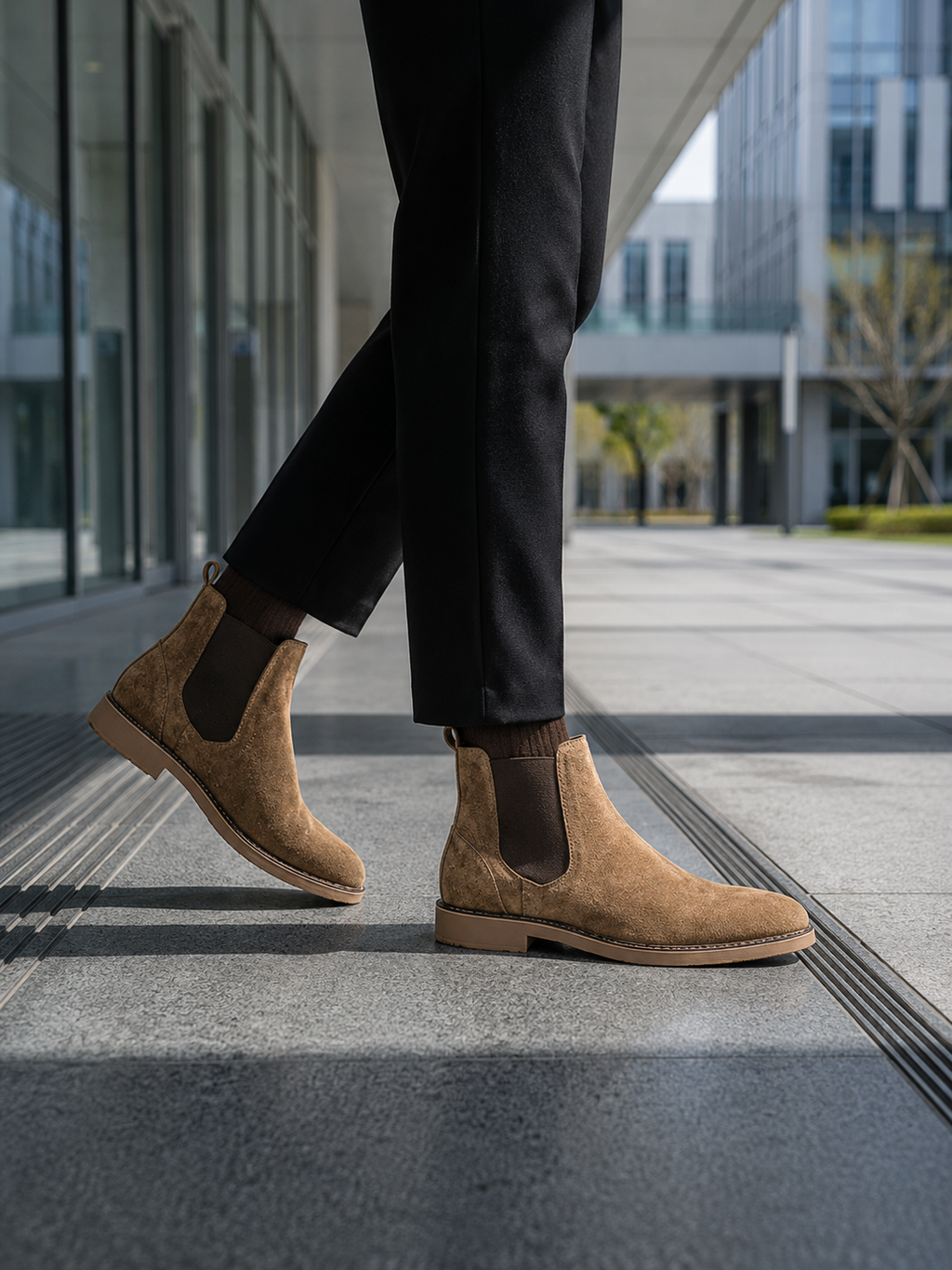 Person wearing brown suede boots and black pants walking on a modern urban street.