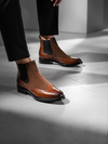 Brown leather Chelsea boots worn with black pants on a gray floor.