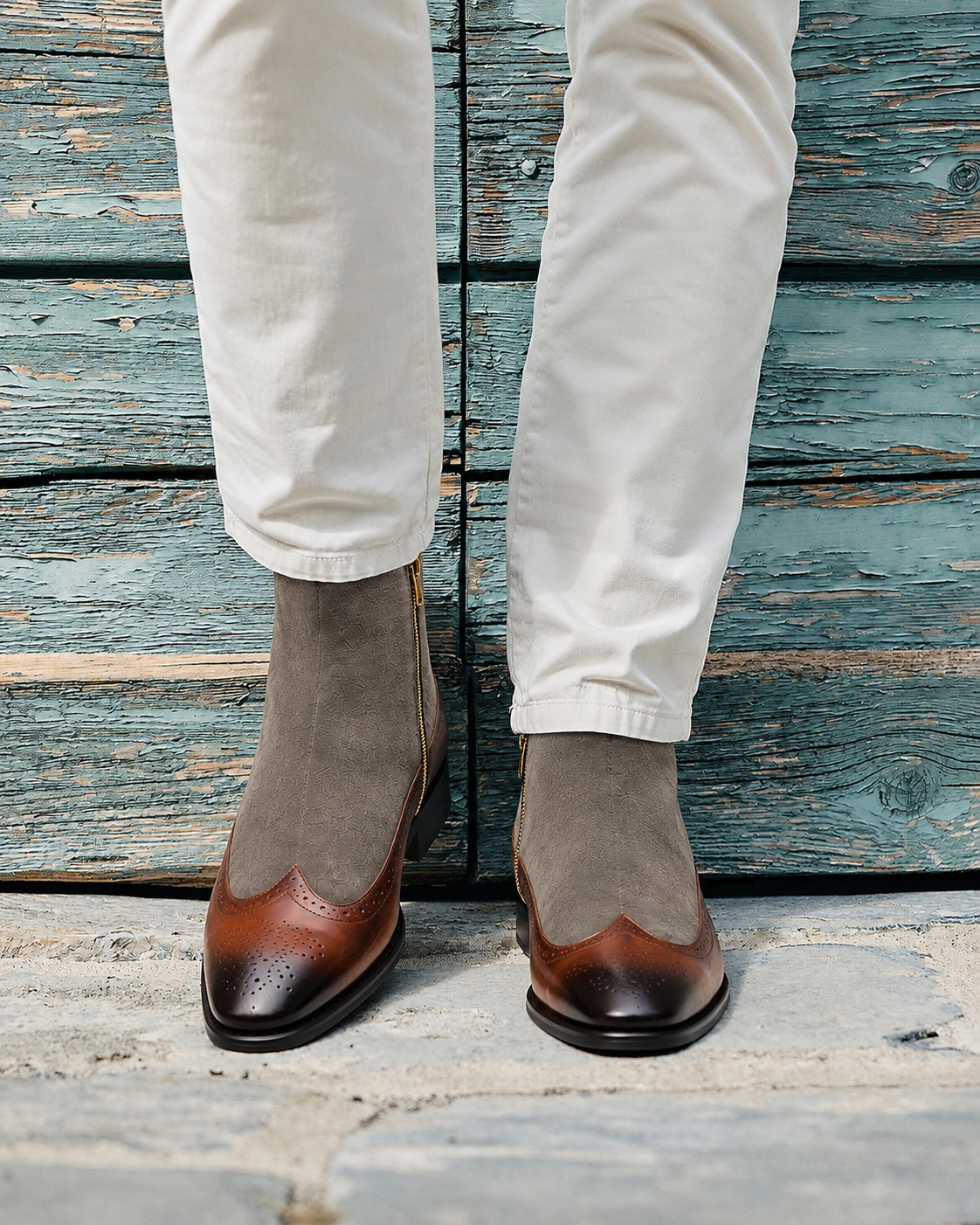 A person wearing off-white trousers and brown suede Chelsea-style wingtip boots with brogue detailing, standing against a rustic teal wooden plank wall.