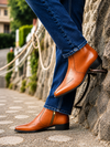 A close-up lifestyle shot of a person wearing blue jeans and tan leather ankle boots with side zippers, standing on a stone path against a rustic stone wall.