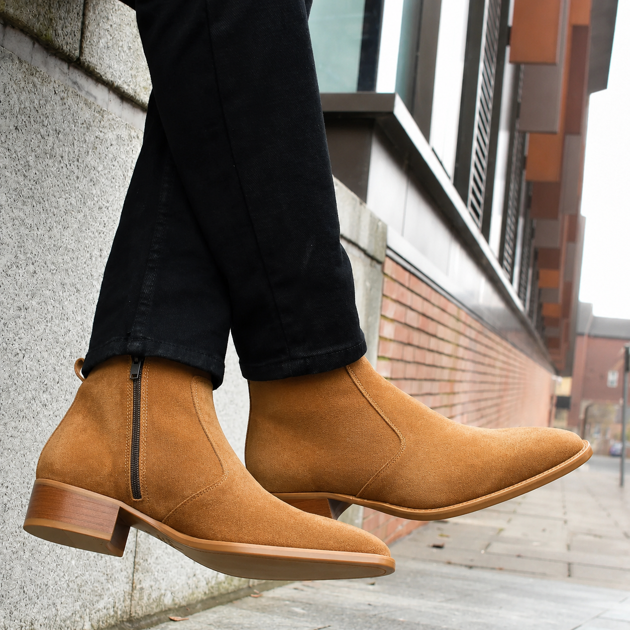 A person wearing tan suede Chelsea-style ankle boots with side zippers and black jeans, photographed outdoors against a modern building wall and sidewalk.